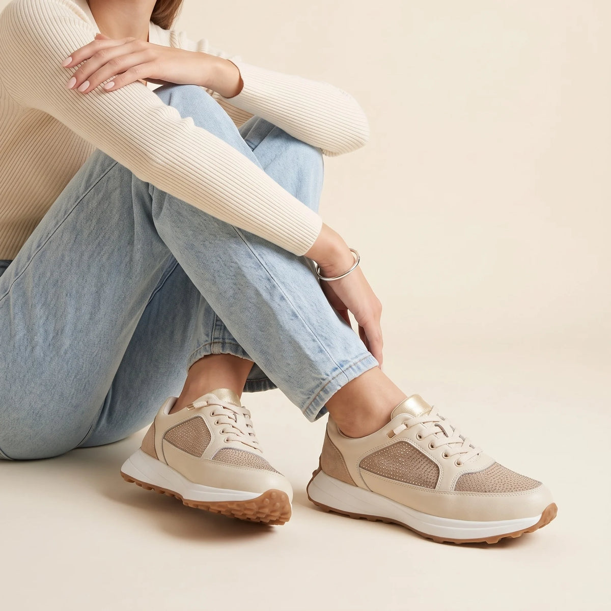 A low-top sneaker in beige and off-white with textured mesh panels, a thick white midsole, and a brown gum outsole
