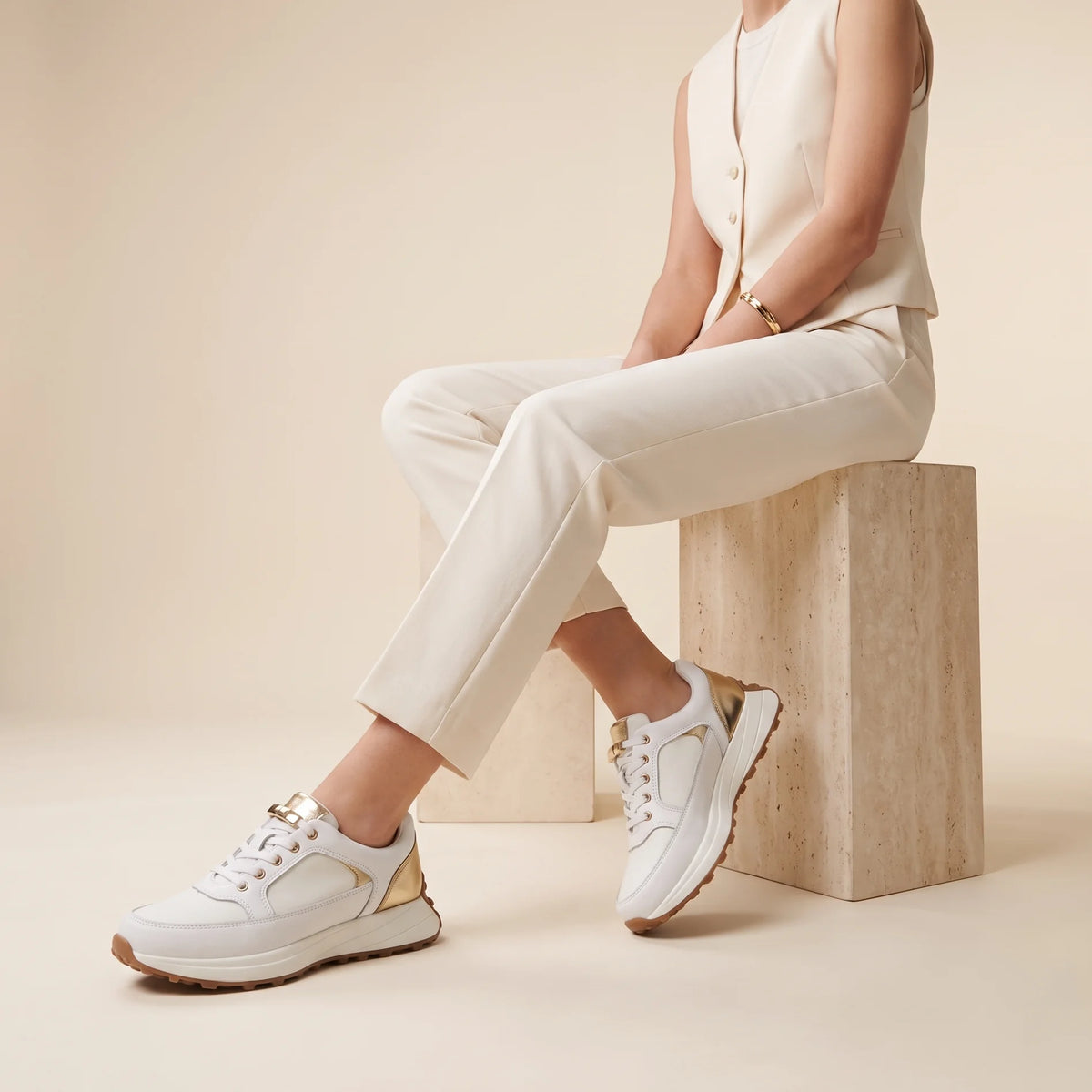 A white low-top sneaker with metallic gold accents on the heel, tongue, and eyelets, featuring a chunky white midsole and a brown gum outsole