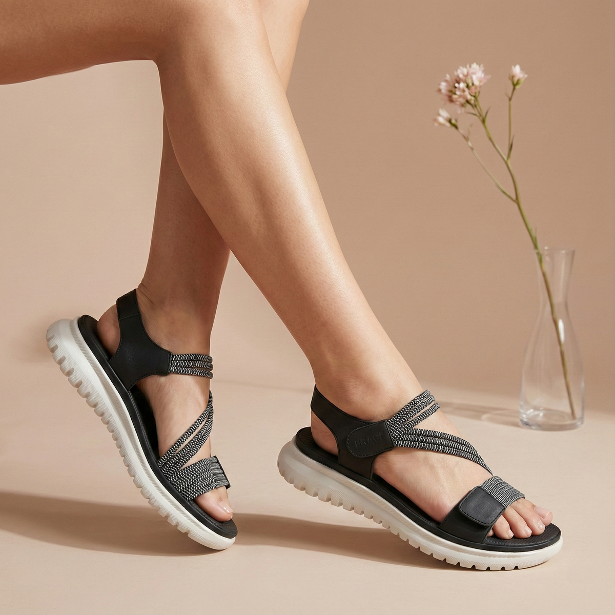 Black sandals worn by a person on a beige background with a vase of flowers.